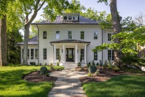 A white house with a tall tree providing shade on the brick pavers walkway that leads up to the front door